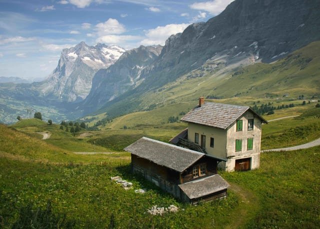 An old stone chalet on a green alpine slope below snowy peaks