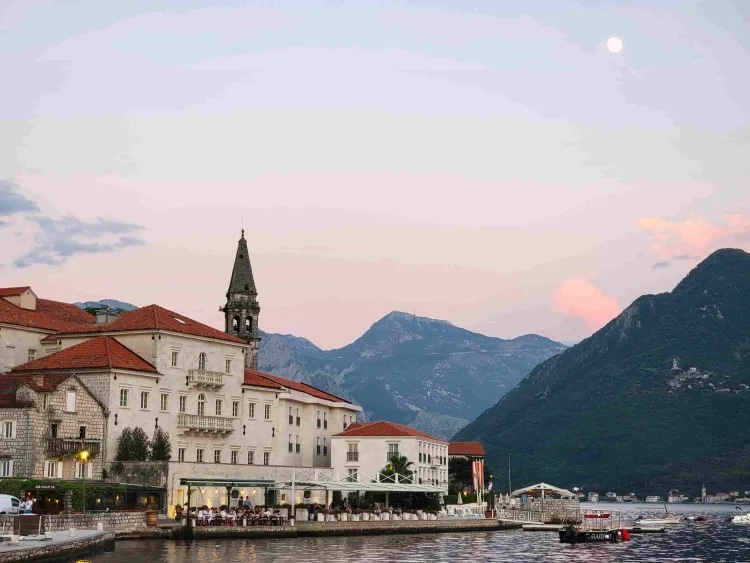 Stone waterfront hotel and bell tower beneath mountains at dusk