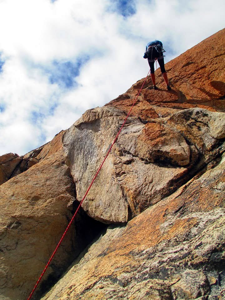 A climber on a rope leading up warm-colored granite against a cloudy sky
