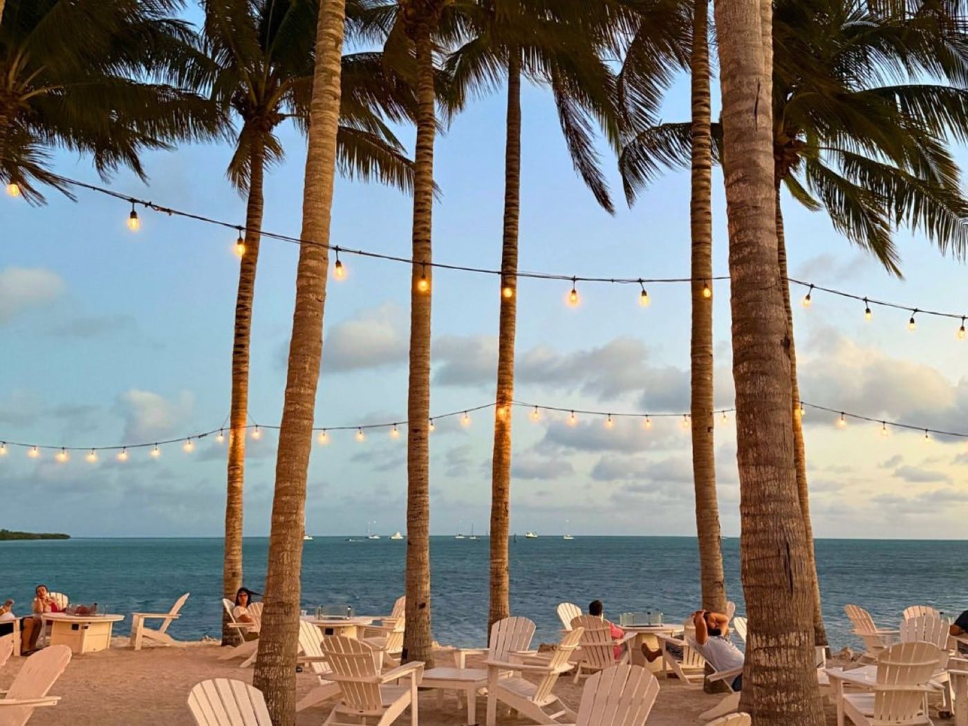 Palm trees strung with lights over white Adirondack chairs on the beach at Isla Bella Beach Resort & Spa